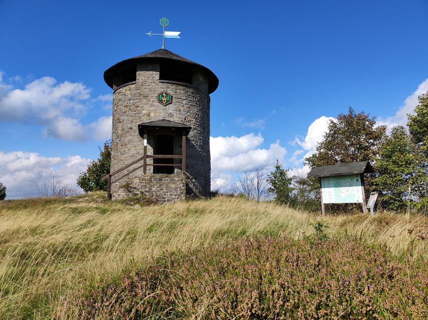 Das Bild zeigt einen runden steinernen Aussichtsturm auf einem grasbewachsenen Hügel vor einem leicht bewölkten Sommerhimmel. Neben dem Aussichtsturm ist eine Informationstafel zu sehen, im Vordergrund Heidekraut.