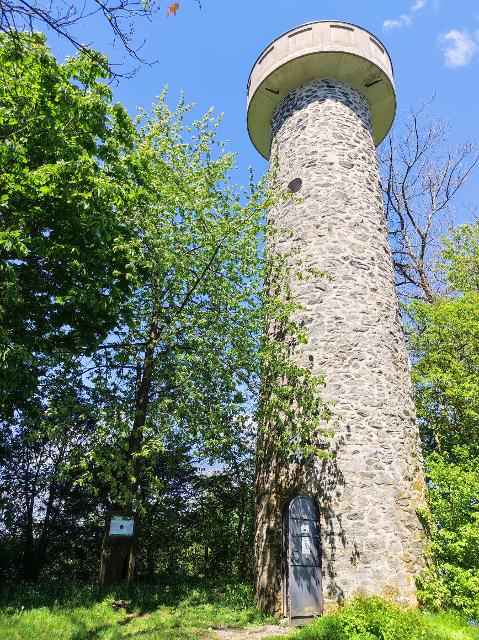 Das Bild zeigt einen runden Turm aus Stein mit geöffneter Stahltür als Eingang. Neben dem Turm sind grüne Büsche und Bäume zu sehen.