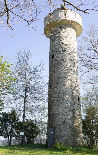 Das Bild zeigt einen steinernen runden Aussichtsturm umgeben von Bäumen unter einem blassblauen Himmel.