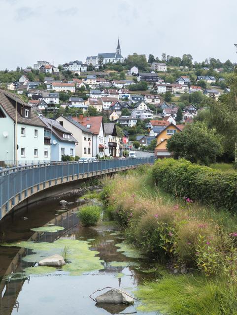 Das Bild zeigt auf der linken Seite einen Fluss. Rechts und im Hintergrund sieht man das Dorf Wilhelmsthal. Ganz oben im Bild sieht man den Kirchturm.