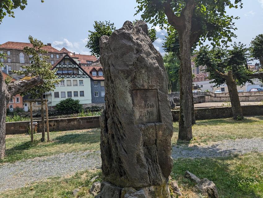 Das Bild zeigt einen Gedenkstein im Vordergrund. Dieser steht in der Jahnsallee in Kronach. Dahinter sieht man Bäume, sowie einen Teil der Altstadt.
