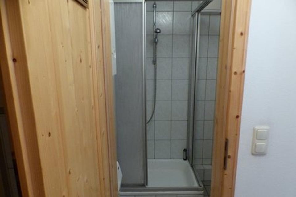 Das Bild zeigt eine Duschkabine, im Vordergrund sind ein Türrahmen und eine geöffnete Holztür zu sehen.