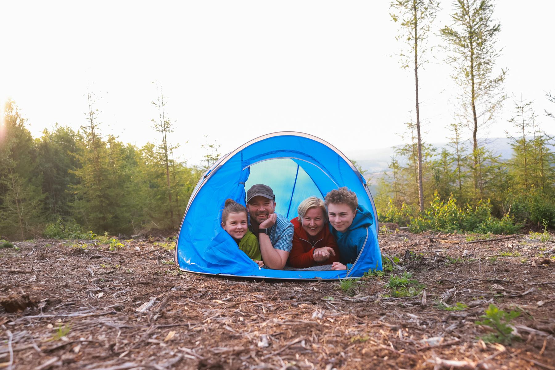 Zwei Kinder und zwei Erwachsene liegen in einem kleinen Zelt. Sie schauen heraus und lächeln. Das Zelt ist blau. Es steht auf einer Fläche, dahinter ist Wald.