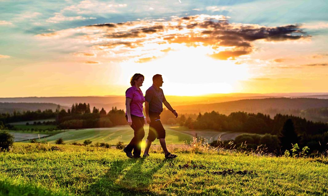 Zwei Personen laufen über eine grüne Wiese. Im Hintergrund geht die Sonne unter. Der Betrachter des Bildes kann weit in die Ferne blicken.