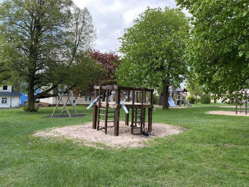 Das Bild zeigt eine Gesamtübersicht des Spielplatzes im Volkspark Helmbrechts. Man sieht eine Schaukel, Kletterturm mit Rutsche und diverse Sandspielmöglichkeiten.
