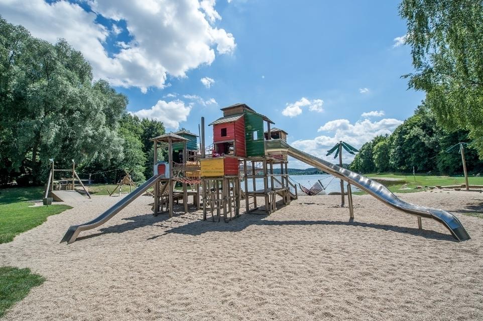 Das Bild zeigt den Spielplatz am Untreusee in Hof. Man sieht eine Seilbahn, Sandspielmöglichkeiten, Hängeschaukel, Kletterturm mit 2 Rutschen.