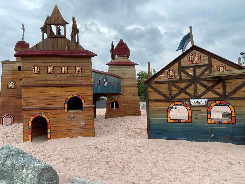 Das Bild zeigt einen Ausschnitz des Burgenspielplatzes. Man sieht die Burg mit unterschiedlichen Tprmen und ein Brauhaus. Diese Gebäude sind mit bunten Fensterrähmen versehen und diesen zum Spielen.
                 title=