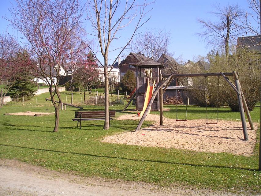 Das Bild zeigt den Spielplatz in Langenbach. Man sieht einen Sandkasten, Schaukel, Wippe und einen Kletterturm mit Rutsche.