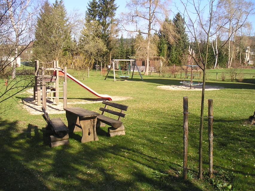 Das Bild zeigt den Spielplatz in Steinbach. Es gibt eine Nestschaukel, einen Kletterturm mit Rutsche, Sitzbänke und ein Klettergerüst.