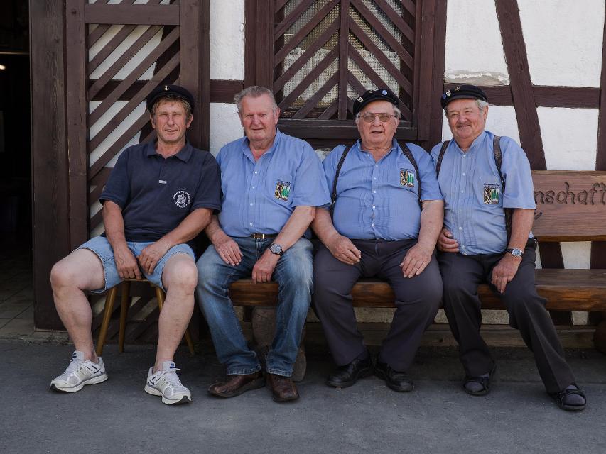 Das Bild zeigt vier Männer auf einer Bank. Sie tragen blaue Hemden und sind als Flößer erkennbar