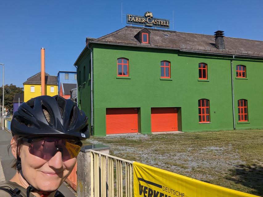 Das Bild zeigt ein grünes Gebäude mit roten Fensterrahmen und davor eine Radfahrerrin, die in die Kamera lächelt.
                 title=