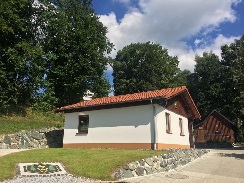 Das Bild zeigt die zwei Hütten auf dem Gelände des Frankenwaldvereins Wartenfels. Im Vordergrund ist ein gepflastertes Logo des Frankenwaldvereins zu sehen, dahinter die gemauerte Hütte. Im Hintergrund sieht man die hölzerne Hütte.