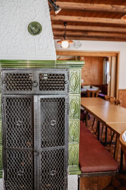 Das Bild zeigt den Kachelofen im Gastraum des Wanderheims Marlesreuth. Im Vordergrund ist die Kachelofentür zu sehen, im Hintergrund Tische und Stühle.