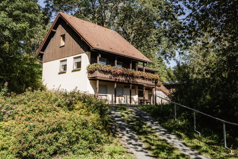 Das Bild zeigt das Wanderheim Marlesreuth im Sommer. Im Vordergrund ist der Aufgang zum Wanderheim sowie ein Metallgeländer zu sehen. Am Balkon des Wanderheims hängen bunte Blumenkästen.