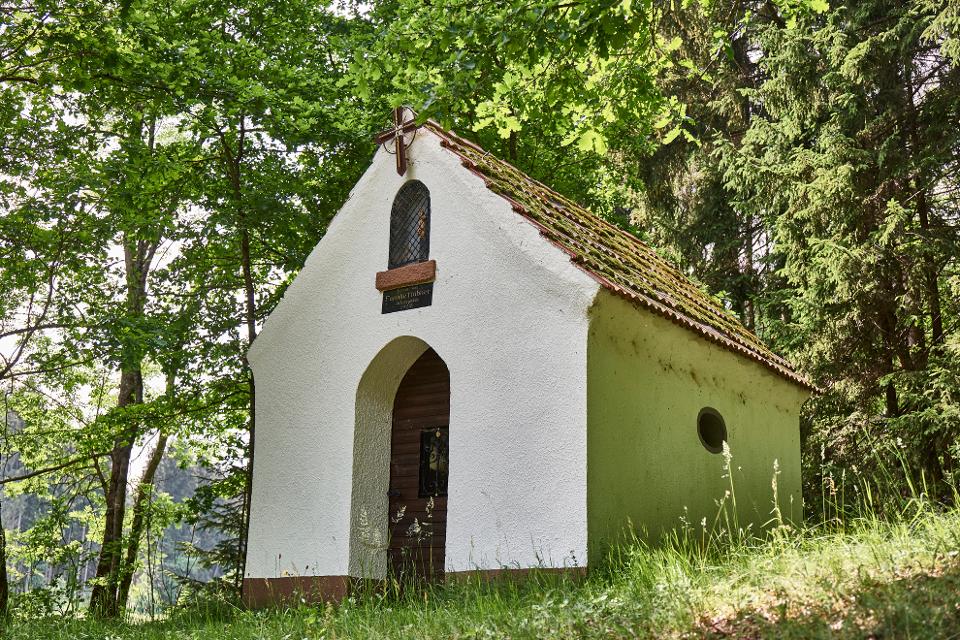 Das Bild zeigt eine kleine weiße Kapelle mit rotem Dach. Im Hintergrund sind grüne Laubbäume zu sehen.
