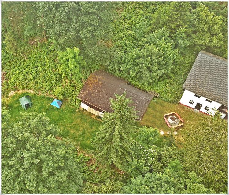 Das Bild zeigt zwei Gebäude, eine Feuerstelle und zwei Zelte aus der Luft, umgeben von grünen Bäumen und grüner Wiese