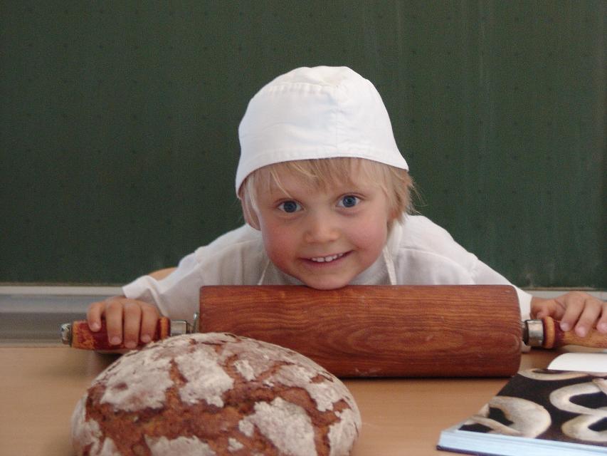 Das Bild zeigt ein Kleinkind in Bäckerkleidung. Es hält einen Teigroller. Vor ihm liegt ein Brotlaib