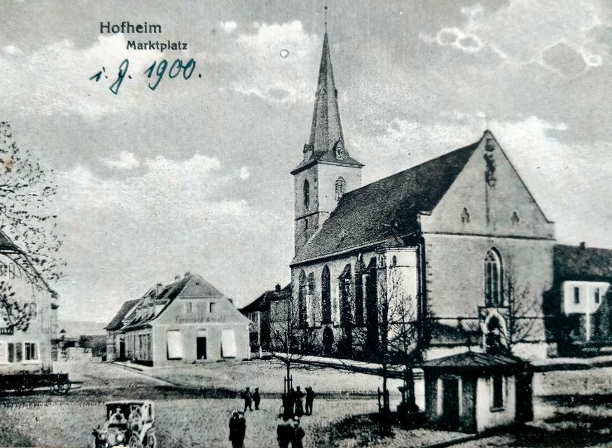 ▶️ Erfahren Sie mehr über die Kirche und warum die Hofheimer Märkte früher meist gar nicht auf dem Marktplatz stattfanden. Das Bild zeigt den Hofheimer Marktplatz um etwa 1900.Sie sehen einen Pumpbrunnen, das Waag...