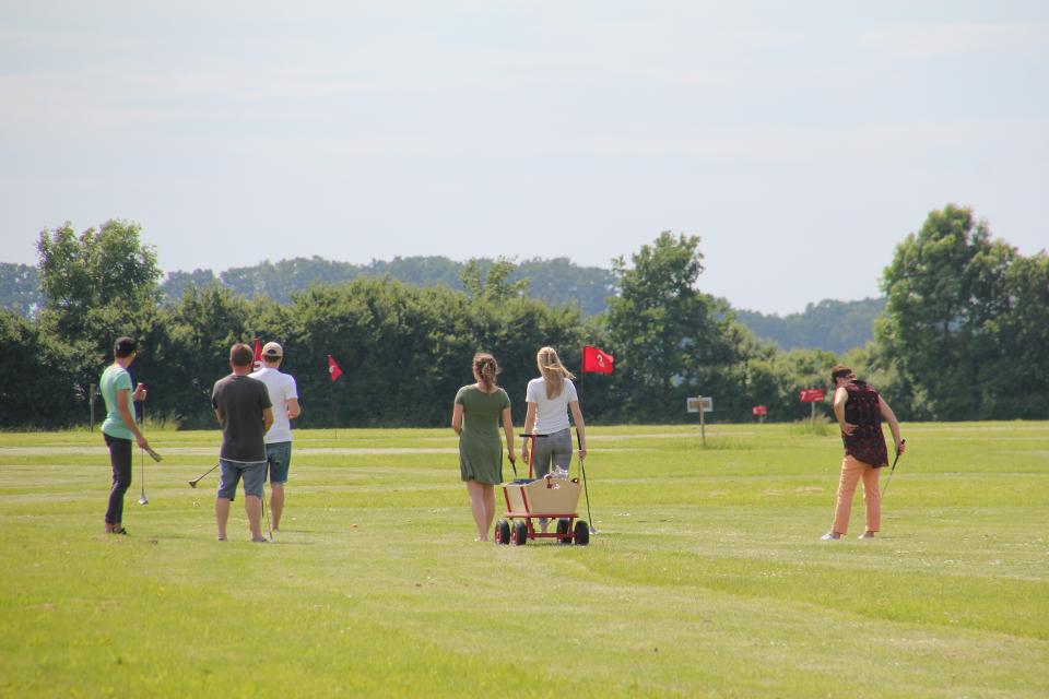 Eine sommerlich gekleidete Gruppe junger Erwachsener steht mit Golfschlägern und einem Bollerwagen auf einem Swin-Golfplatz auf dem rote Fähnchen die zu spielenden Löcher markieren.