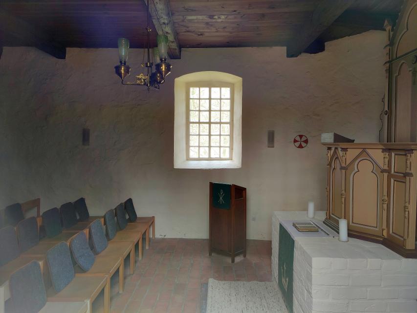 Schlicht gestalteter Altarraum der St. Michaelis-Kapelle in Müssingen mit mehreren Sitzreihen, Altar, Pult, an die Wand gemaltem Weihekreuz und einem Fenster.