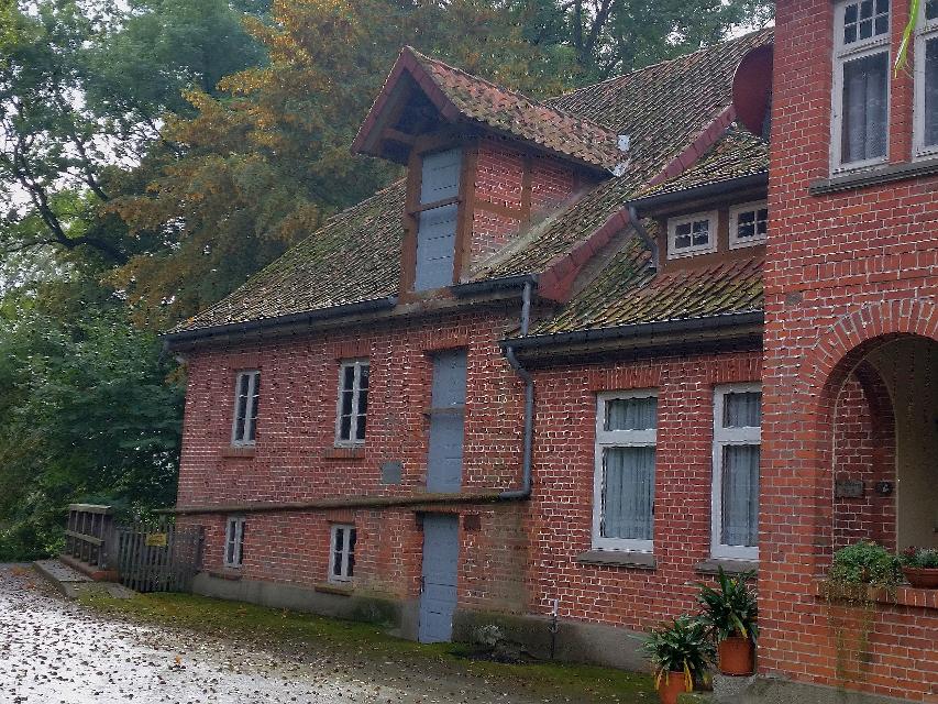 Blick auf das Gebäude der stillgelegten Wassermühle Kroetzmühle, ein dreigeschossiger Backsteinbau mit großen, blau gestrichenen Holztüren in allen Etagen. Davor die regennasse Straße und etwas Herbstlaub.