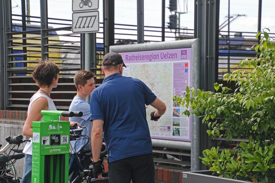 3 Radfahrer stehen vor der Rasreiseregion Uelzen Karte und der Fahrrareparaturstation am Hundertwasser Bahnhof Uelzen.