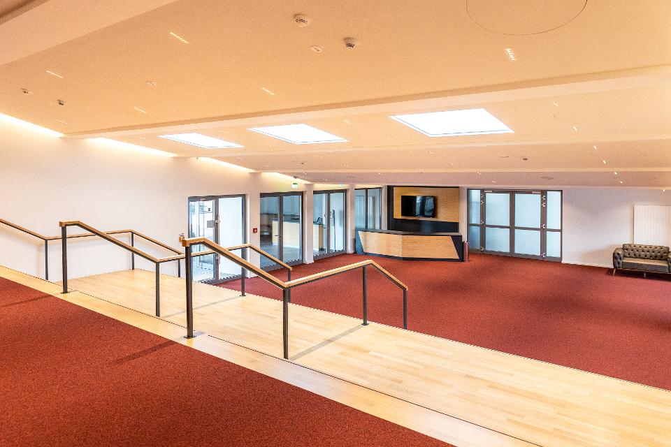 Zu sehen ist das Foyer des Theaters an der Ilmenau. In der hinteren Ecke ist ein Tresen mit Bildschirmen. Im Vordergrund Treppen mit Geländer.