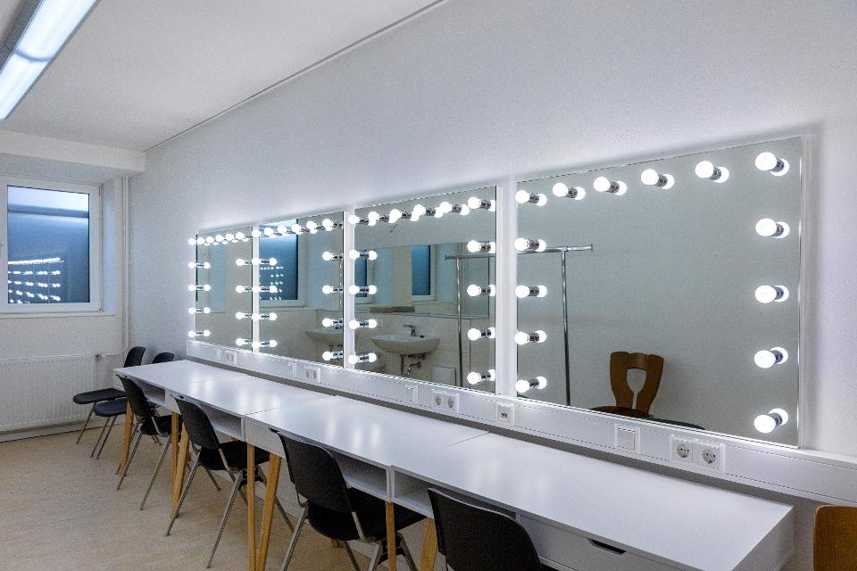 Zu sehen ist die Garderobe im Backstagebereichs des Theaters an der Ilmenau. Ein langer Tisch mit Stühlen steht vor einer Front aus vier Spiegeln mit integrierten Leuchten.