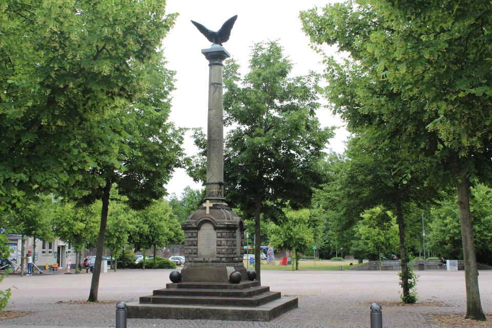 Kriegerdenkmal Säule mit fliegendem Adler auf der Spitze. Der untere Teil des Denkmals besteht aus Stufen und ist versehen mit einem Kreuz und einem Schriftzug.