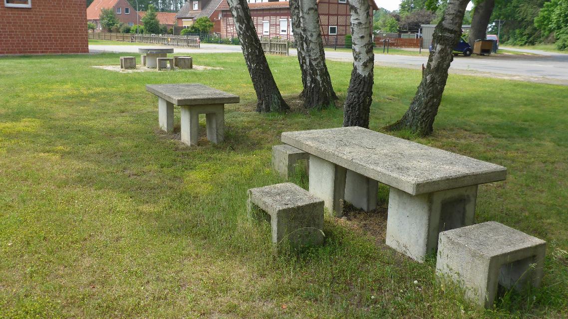 Zwei aus Beton gefertigte Tische und mehrere Betonhocker stehen auf einer Wiese unter Bäumen