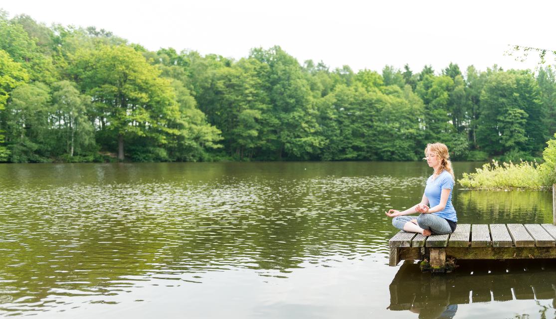 Frau sitzt auf einem Steg am See und meditiert, im Hintergrund Bäume