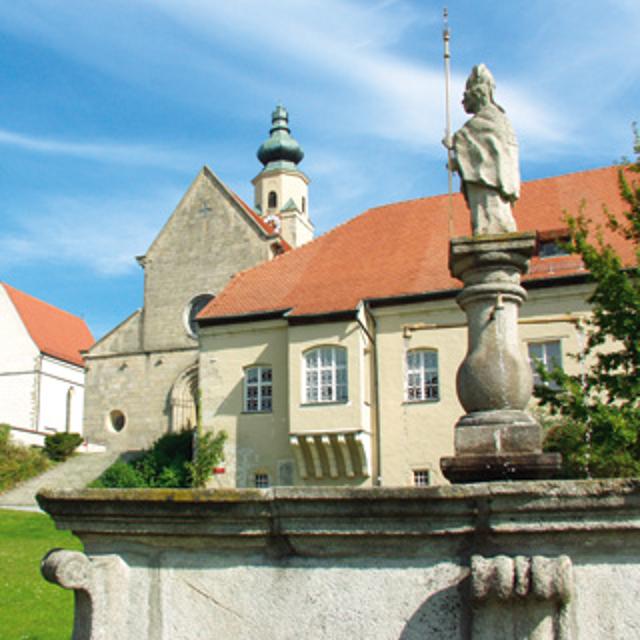 Teilansicht der Klosterkirche Windberg von außen. Im Vordergrund steht auf einer verschnörkelten Mauer die Statue des Ordensgründers, des Heiligen Norbert.