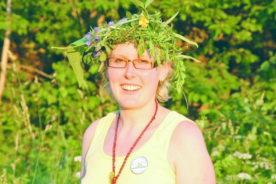 Vor einem Hintergrund aus grünen Blättern steht die sommerlich gekleidete Wanderführerin. Sie trägt einen Kranz aus Wildkräutern auf dem Kopf und lacht fröhlich in die Kamera.