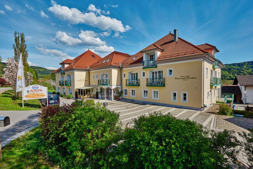 Außenaufnahme des Hotels Bayerwald-Residenz mit grünen Balkonen und Parkplätzen direkt am Haus. Die Hotelzufahrt liegt zwischen blühenden Sträuchern.