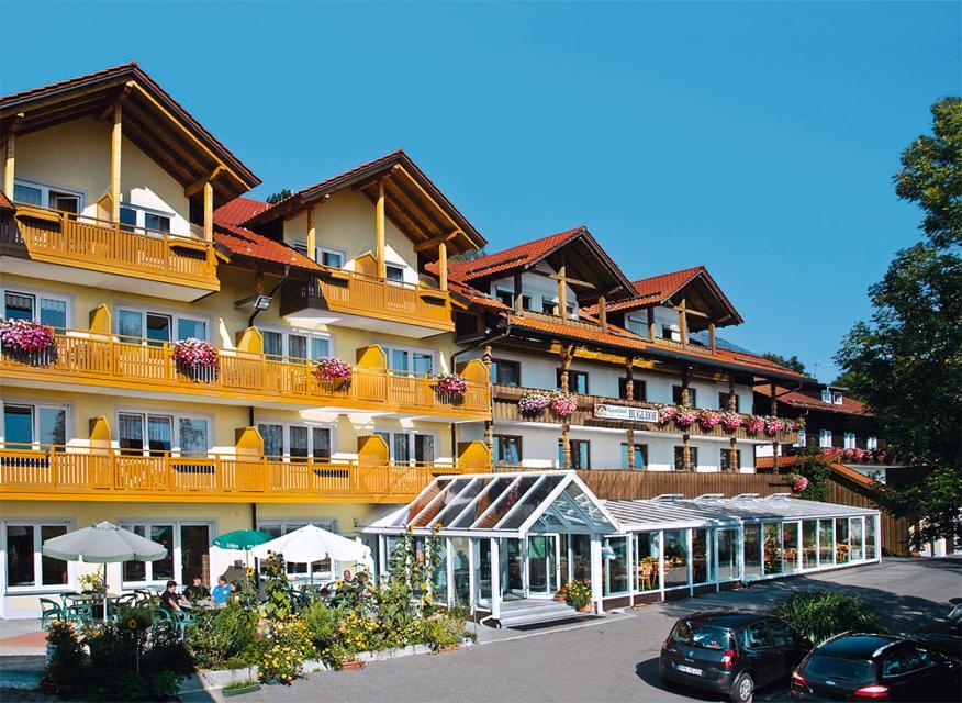 Außenaufnahme vom Gasthof Buglhof mit Wintergarten und sonnenbeschirmter Terrasse. Die Balkone sind blumengeschmückt. Direkt vorm Haus drei parkende Autos.