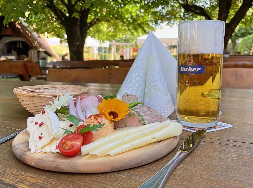 Brotzeitbrett mit verschiedenem Käse, mit Tomaten und Blüten garniert, ein halber Liter Bier. Im Hintergrund Biergarten
