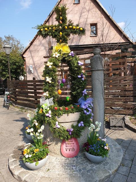 Mit Tannengirlanden, Ostereiern und Blumen geschmückter Brunnen, Krone aus Tannenzweigen mit einem Kreuz an der Spitze