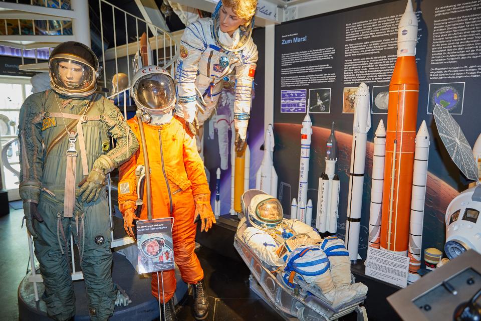 Vier Figuren in unterschiedlichen Raumanzügen neben mehreren Raketenmodellen
