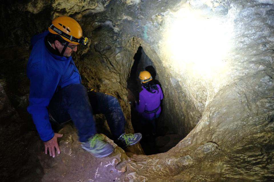 Frau mit Helm geht durch schmalen Spalt in der Höhle, Mann steigt zu ihr hinab