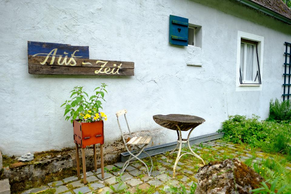 Holzschild "Aus-Zeit" an alter Mühle, davor Blumenkasten mit gelben Blüten und ein Klappstuhl vor einem runden Tischchen auf Pflaster 