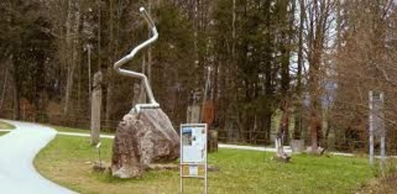 Der Betrachter blickt auf eine Kunstinstallation die einen Stein zeigt auf dem ein Metallrohr angebracht ist das sich nach oben schlängelt. Das Ganze steht auf einem kleinen Stück Wiese, dahinter Wald.
