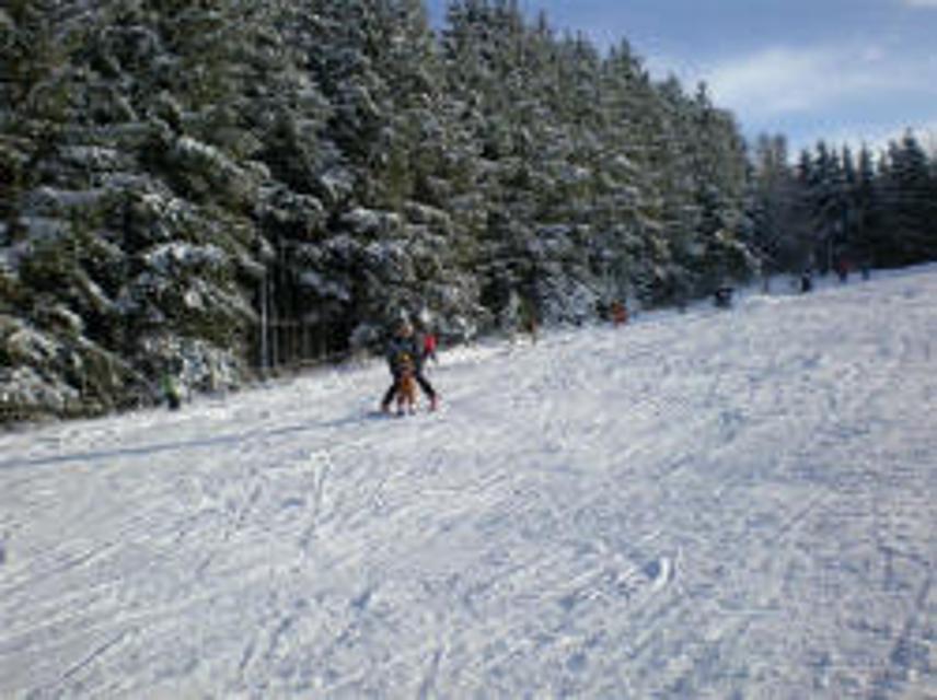 Der Betrachter sieht wie zwei Kinder mit einem Bügellift einen sanften Hang hinaufgezogen werden um Skifahren zu üben.