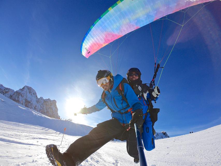 Der Betrachter beobachtet wie zwei Personen im Tandemflug mit dem Gleitschirm auf der geschlossenen Schneedecke landen.