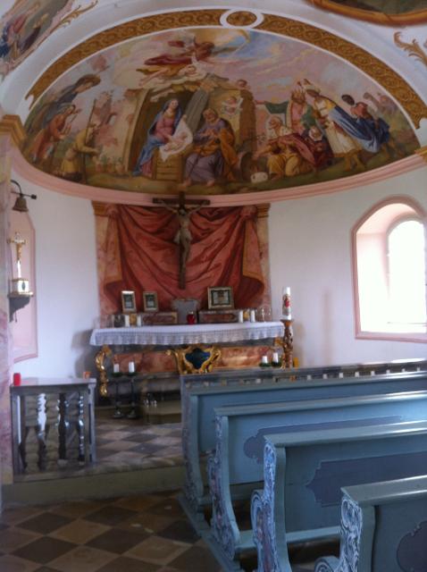 Der Besucher steht in der Kapelle und blickt an hellblauen barocken Kirchenbänken in den Altarraum über welchem sich ein prunkvolles Deckenfresko in der Kuppe befindet.