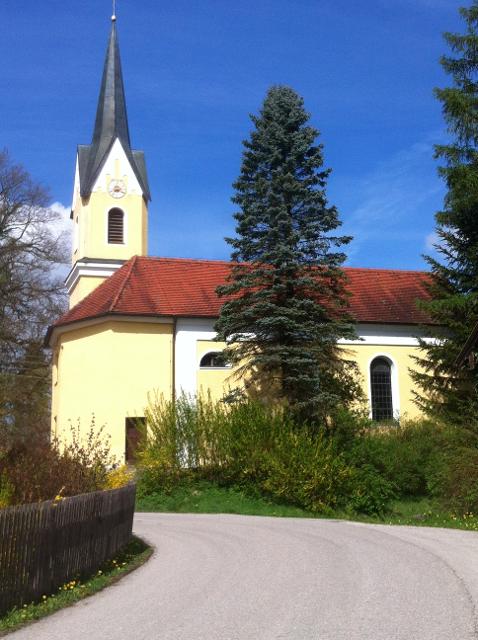 Der Betrachter blickt vor sich auf eine sanftgelbe Kirche die an der Landstraße steht und von ein paar Bäumen und Sträucher eingewachsen ist.