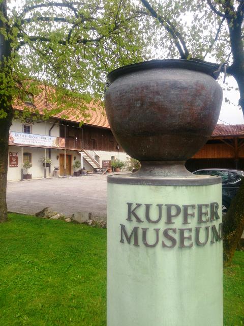 Der Betrachter steht vor einer Säule auf der ein Kupfergefäß angebracht ist mit der Inschrift "Kupfermuseum". Im Hintergrund das Museumsgebäude.