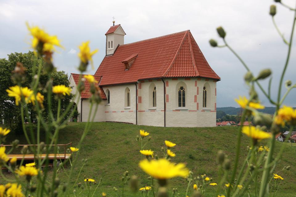 Der Betrachter blickt über eine Blumenwiese hin zu einer relativ großen Kapelle auf einer kleinen Anhöhe.