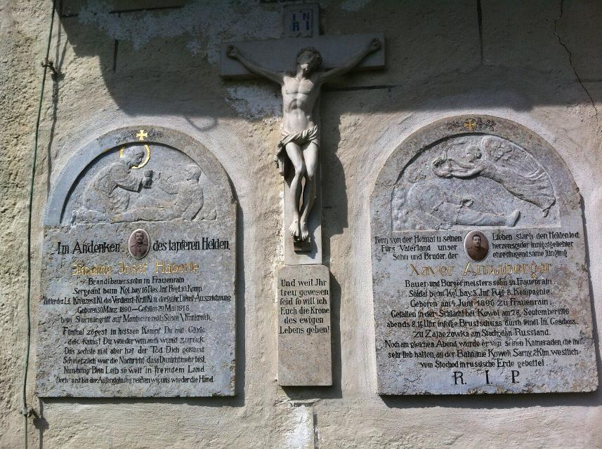 Der Betrachter blickt auf zwei steinerne Gedenktafeln und einem steinernen Kreuz in der Mitte, welche an einer Kirchenwand außen eingelassen wurde.