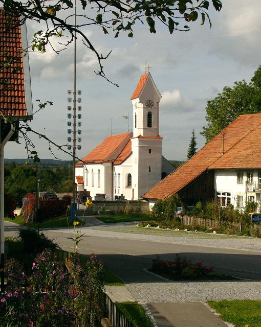 Der Betrachter blickt durch die Dorfstraße auf eine stattliche weiße Kirche und den Maibaum der links davon steht.