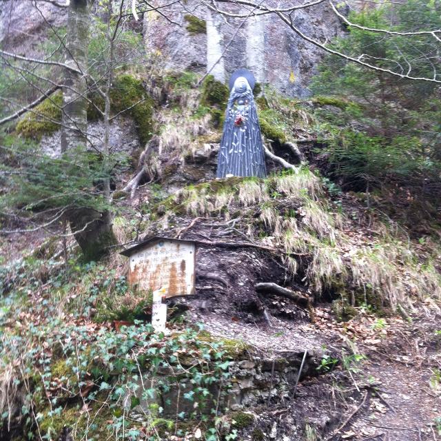 Der Betrachter steht an einem steilen Hang und blickt ein paar Meter hoch wo eine Madonna vor Felsen an einem kleinen Grasvorsprung steht und betet. Ein Naturweg führt an ihr vorbei.